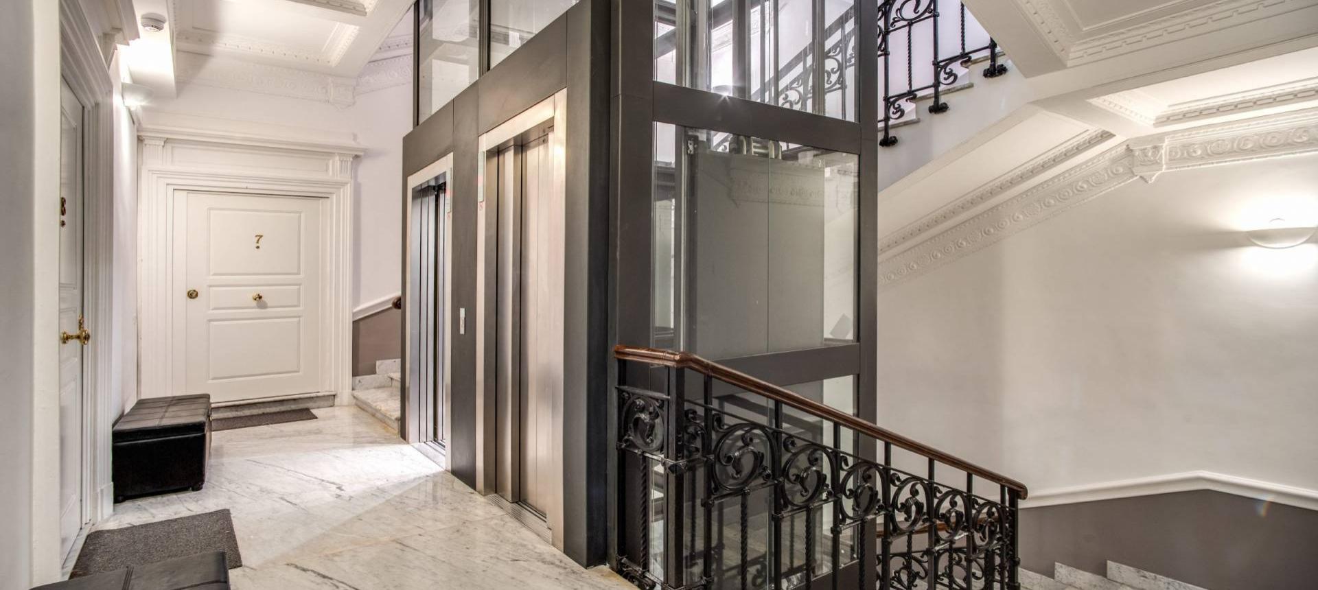 Elegante ingresso con ascensore in vetro e scala in marmo con ringhiera decorata.