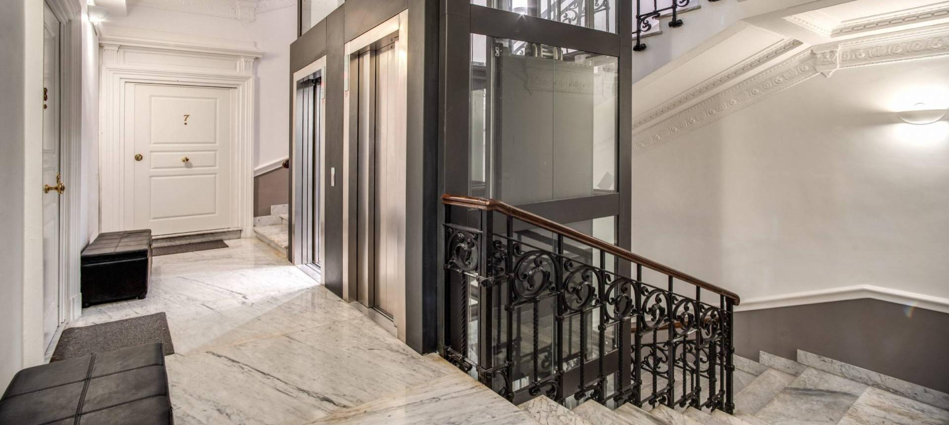 Elegante ingresso con ascensore moderno e scala in marmo, decorazioni classiche e porta bianca.