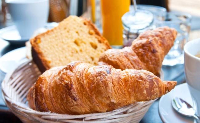 Colazione con croissant, pane, succo d'arancia e caffè in tazza bianca.